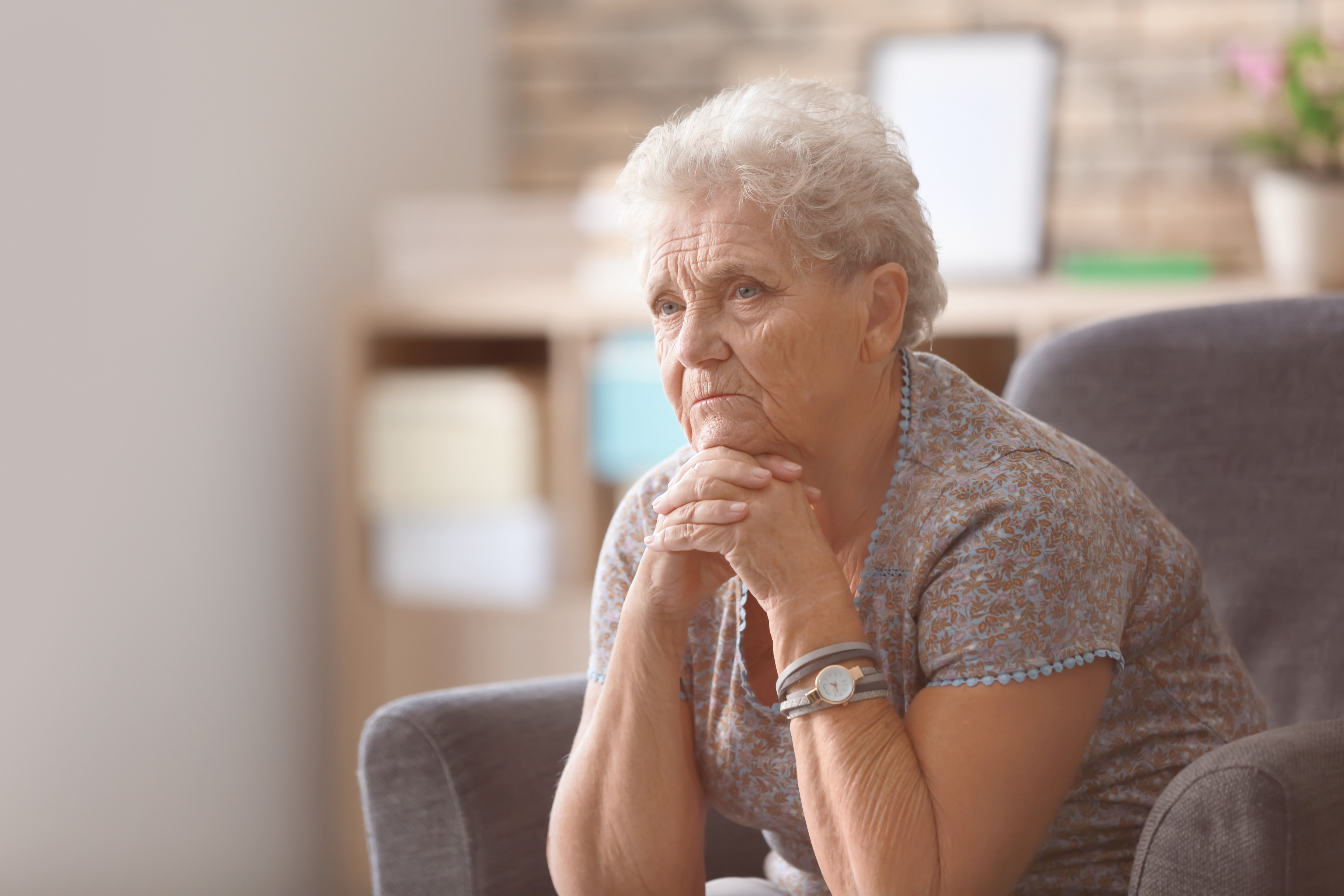 Elderly person with short gray hair, wearing a floral top and a watch, resting chin on hands and looking thoughtful while sitting indoors.